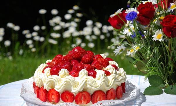 Gräddtårta med jordgubbar på ett bord ute i en trädgård. Bredvid tårtan står en vacker blombukett. 
