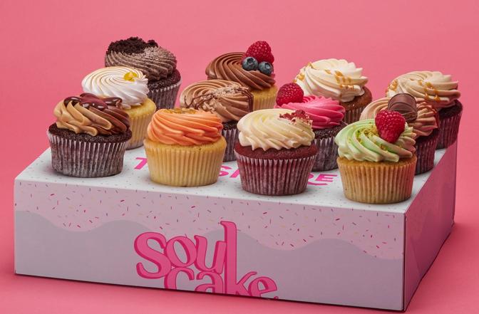 En eske står på en rosa overflate med 12 cupcakes i ulike farger og smaker plassert pent i rader oppå seg. 