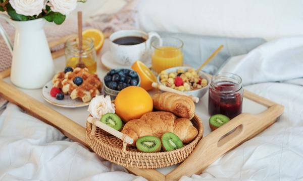 En person som äter frukost på sängen. Personen håller i en kaffekopp och har en bricka full med crossainter, juice, frukt och blommor. 