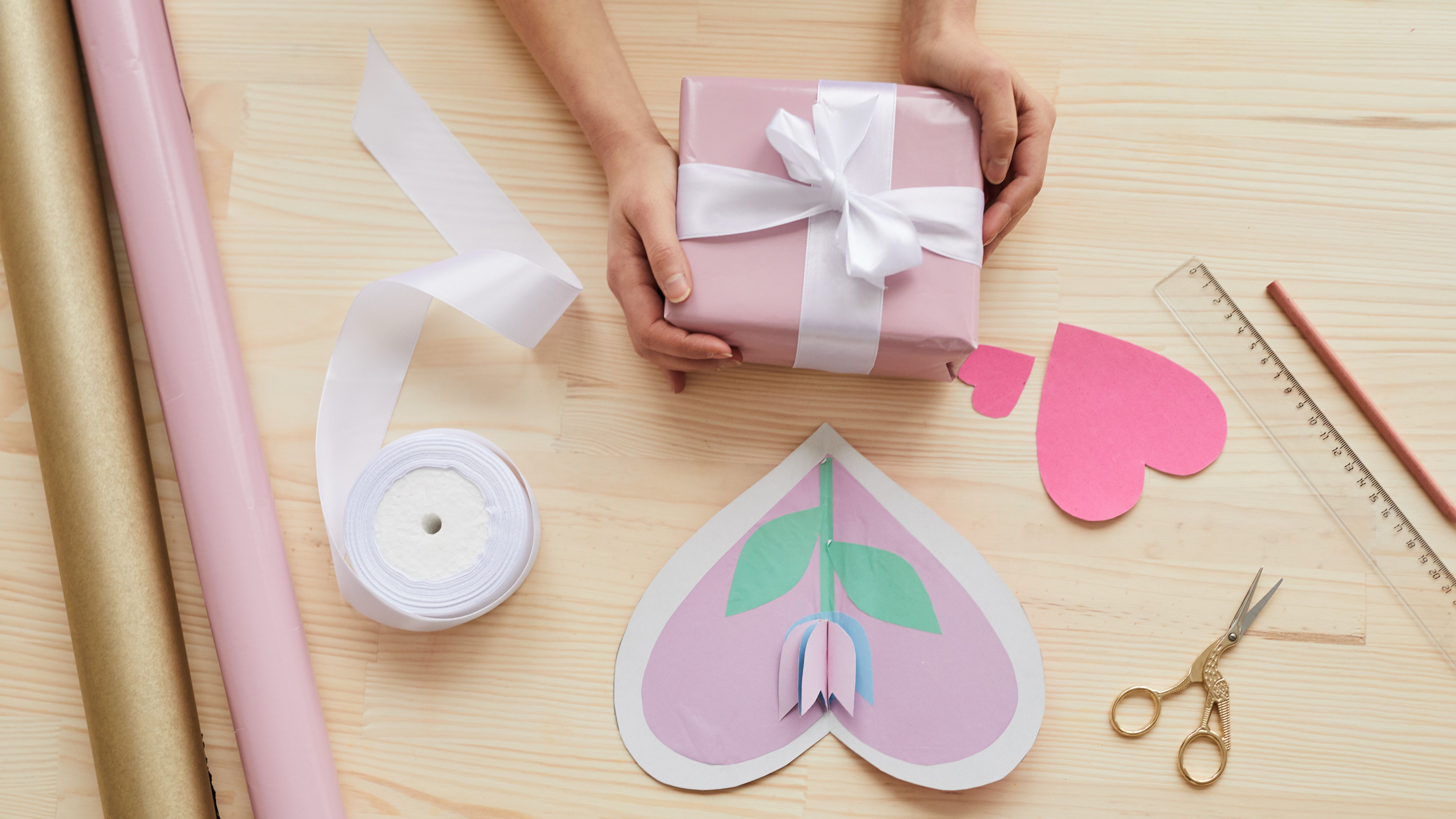 En rosa present med vitt presentsnöre. Omringat av presentpapper, ett hemmagjort kort, sax och linjal. 