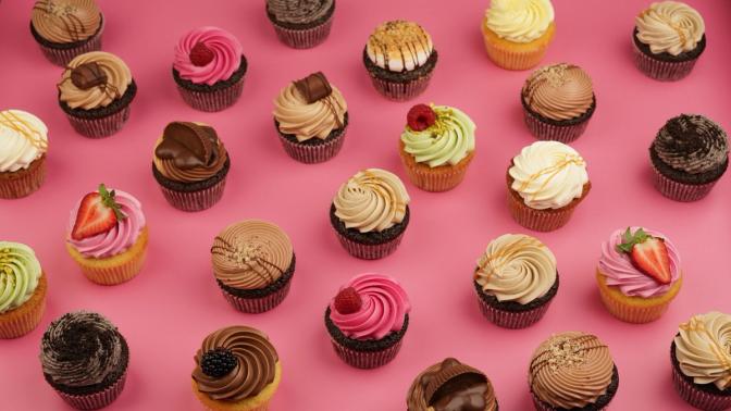Cupcakes i ulike smaker og farger står tilfeldig plassert på en rose overflate. 
