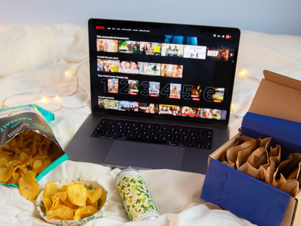 Dator med en streamingtjänst uppe. Datorn står på en säng och är omringad av snacks, till exempel chips och kakor. 