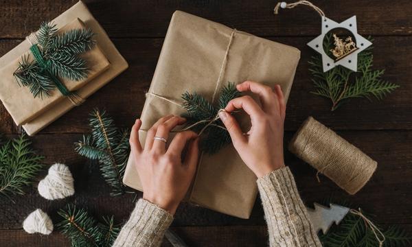 Hender som pakker inn julegave i gråpapir med hyssing og grønne kvister. 