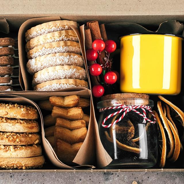 En låda med hembakta kakor, en mugg samt en burk med torkad frukt. Lådan är omringad av granris, pepparkakor och röda bär.