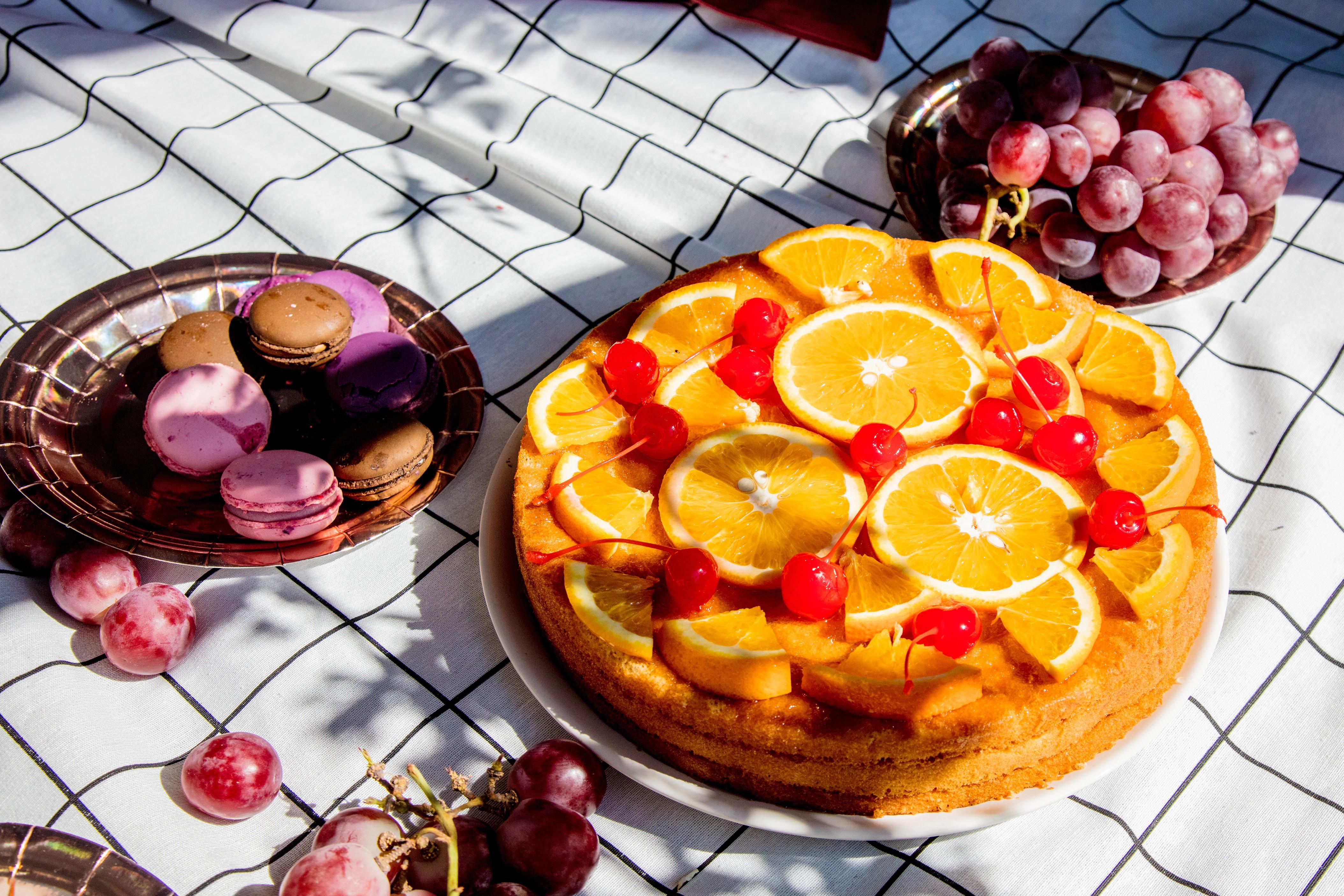 En härlig tårta toppad med apelsinskivor och körsbär. Tårtan står på en picknickfilt och bredvid står en tallrik med vindruvor samt en tallrik med macarons. 