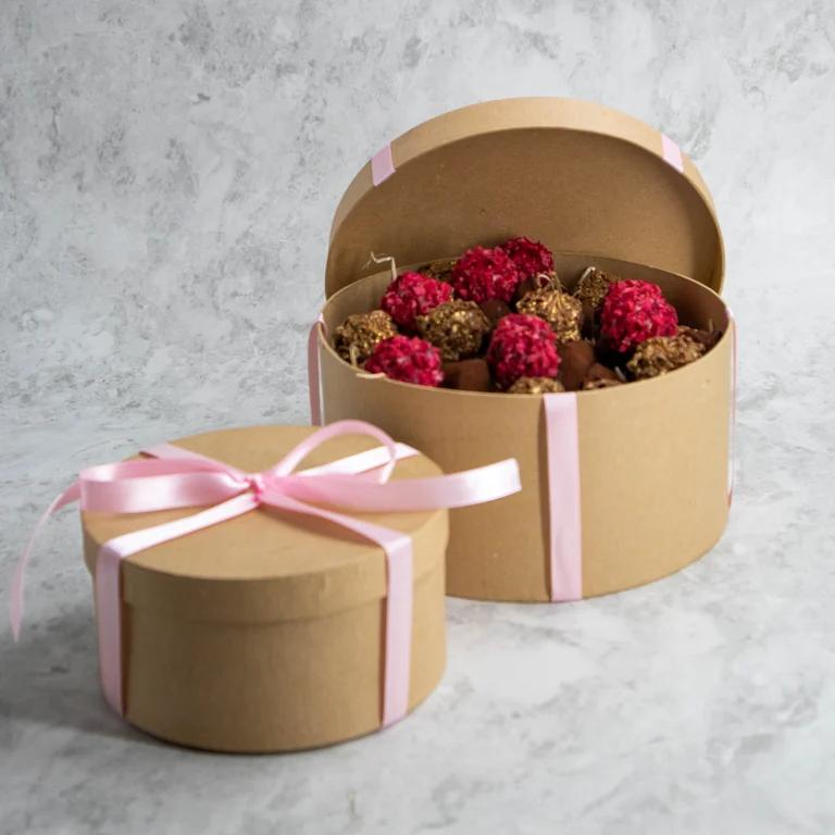 Två runda, ljusbruna lådor som är fulla med praliner i olika smaker. Runt Lådorna är dekorativa, rosa snören knutna. 