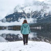 A brown haired woman standing lakeside in front of a mountain range