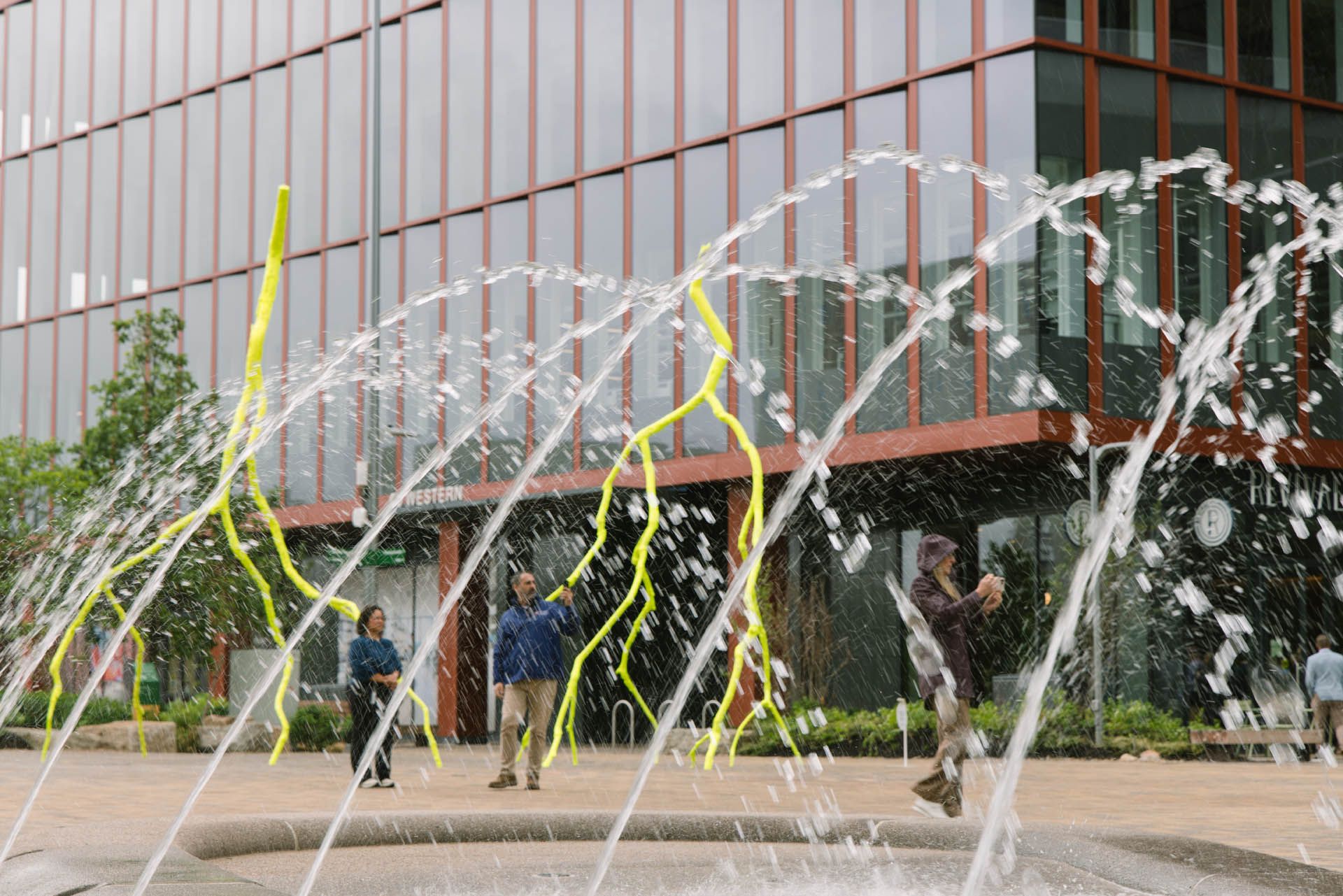 This image shows the newly opened Enterprise Research Campus by Harvard University in Allston, Massachusetts, featuring modern architectural buildings designed by Studio Gang and Henning Larsen alongside a public water fountain plaza.