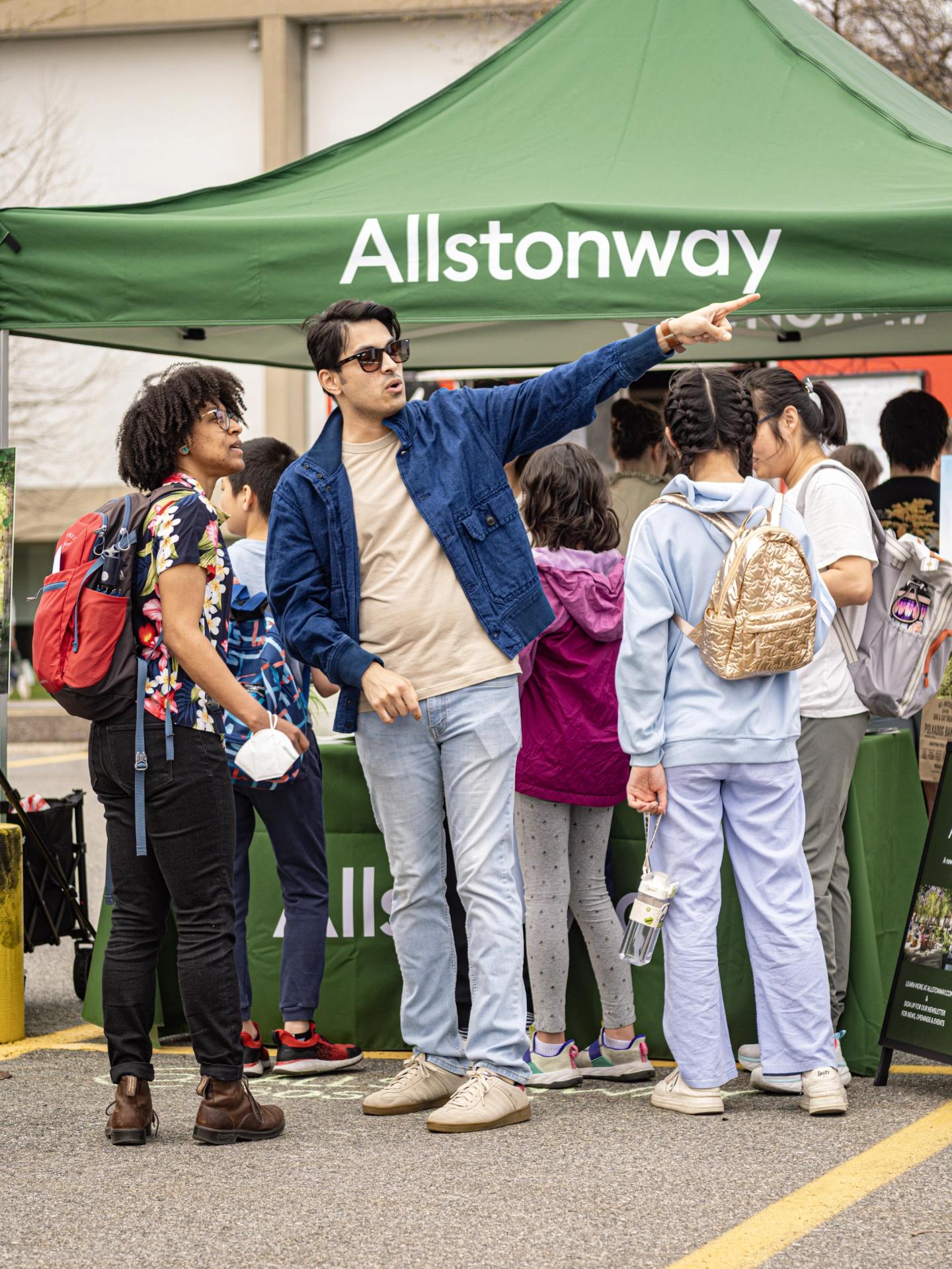 a community gathering with people interacting around a branded booth for "Allstonway," which is a public space project aimed at connecting the community. 
