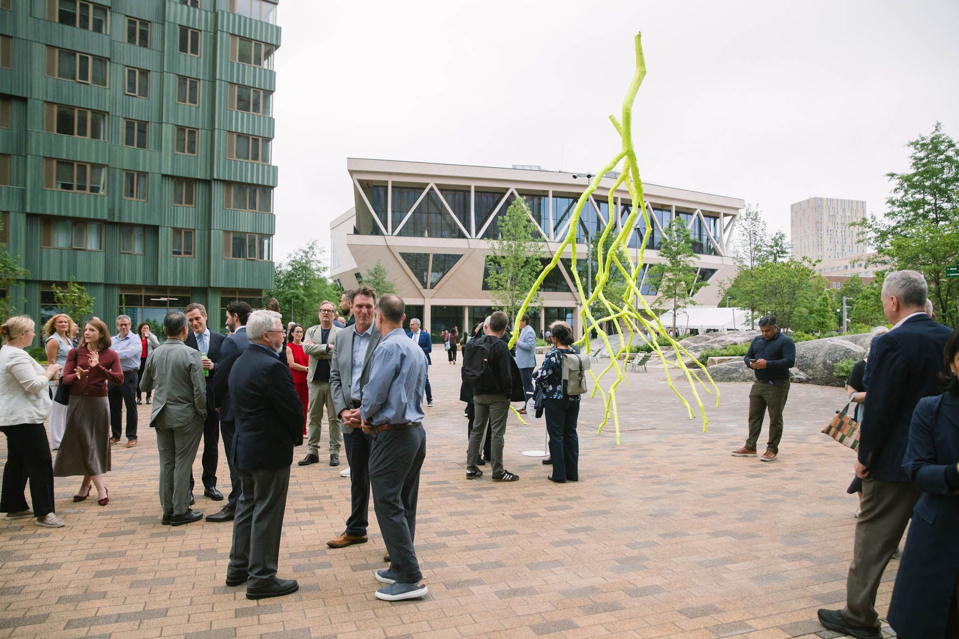 The image features Harvard University's David Rubenstein Treehouse designed by Studio Gang at the Enterprise Research Campus in Allston, Boston. People gather on the paved plaza outside the 55,000-square-foot mass-timber conference center next to a yellow structural art installation.
