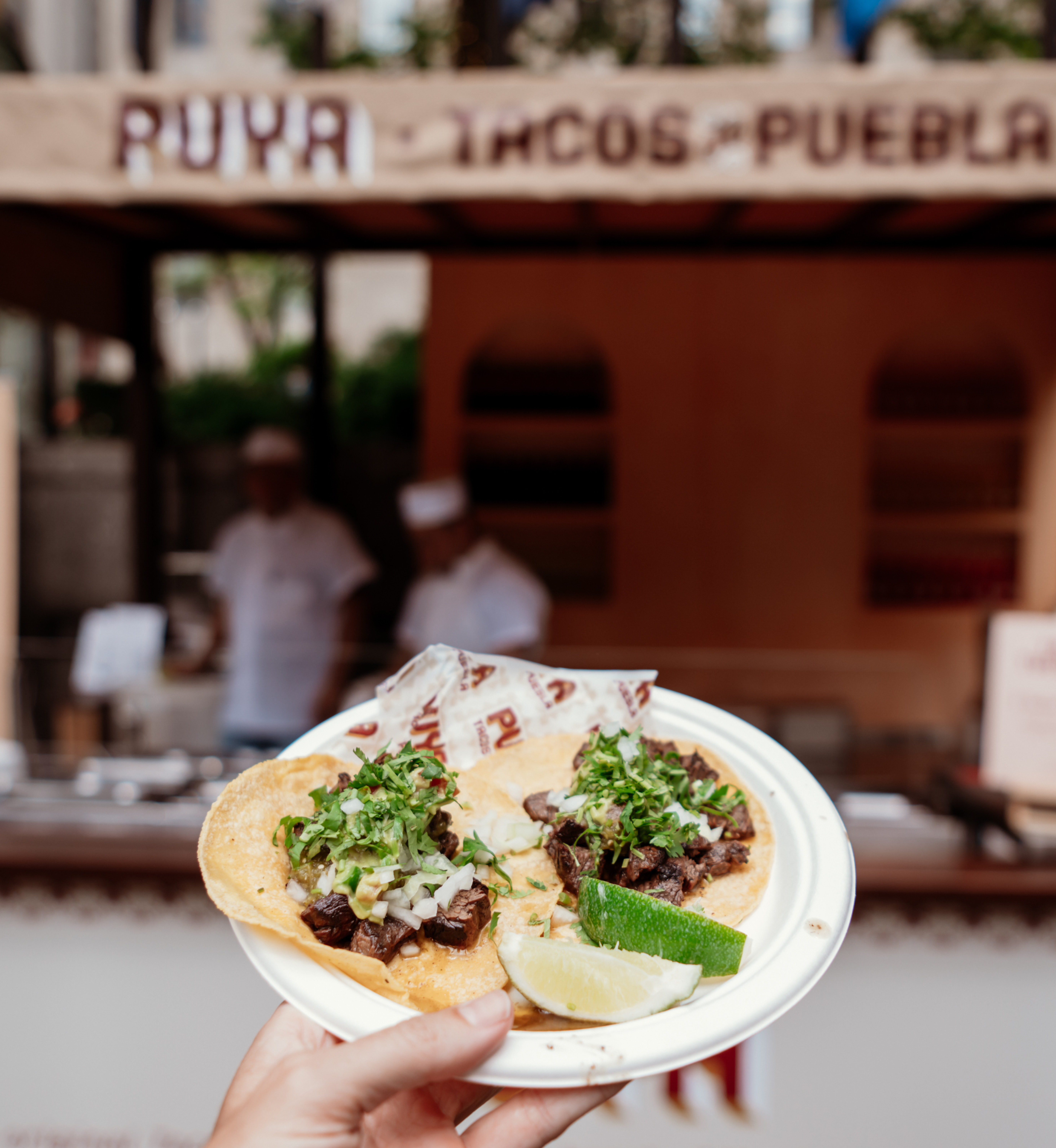 Hand holding a plate of tacos in front of Puya's food truck at Rockefeller Center