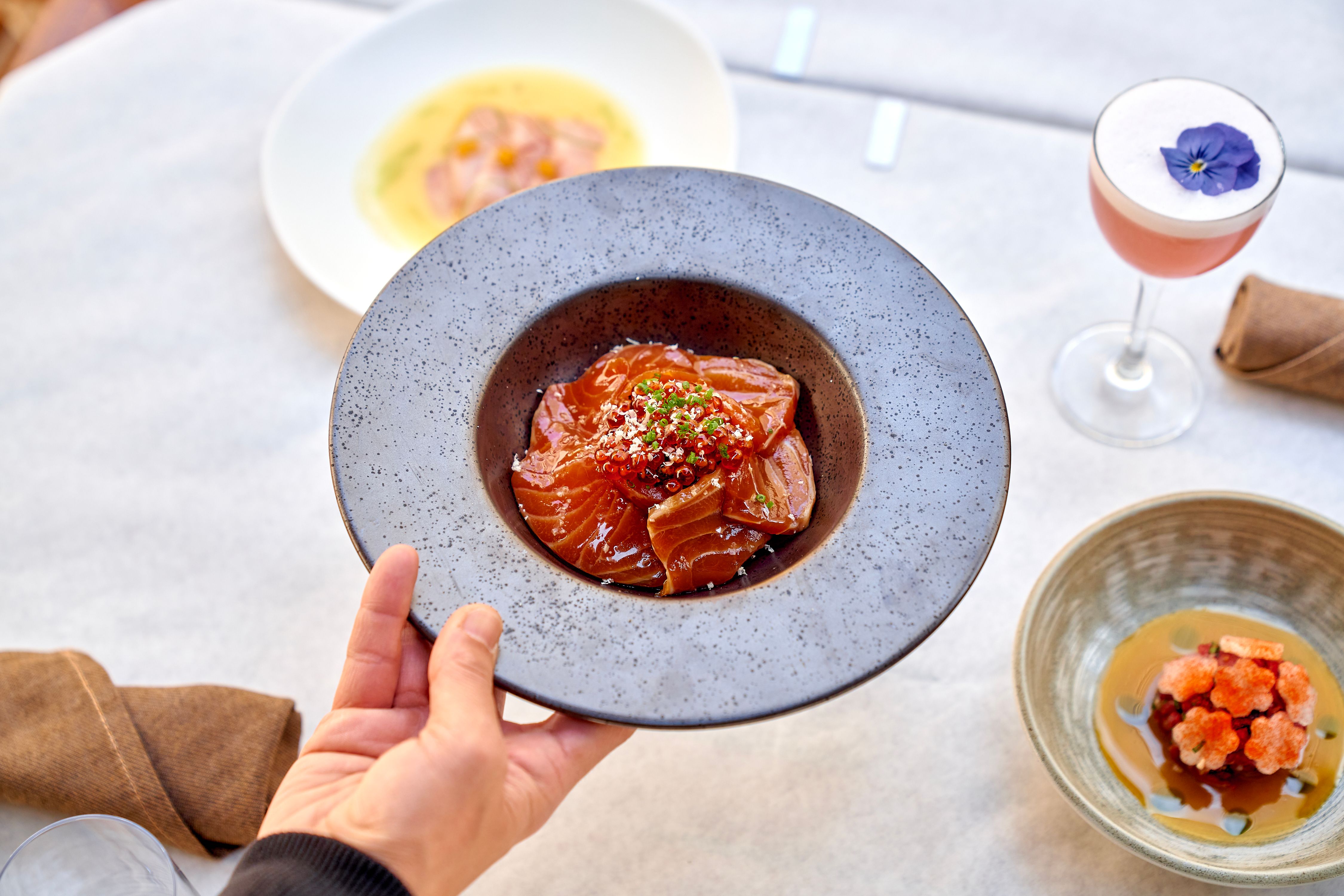 Holding a plate of tuna tartare over a white tablecloth with a pink cocktail
