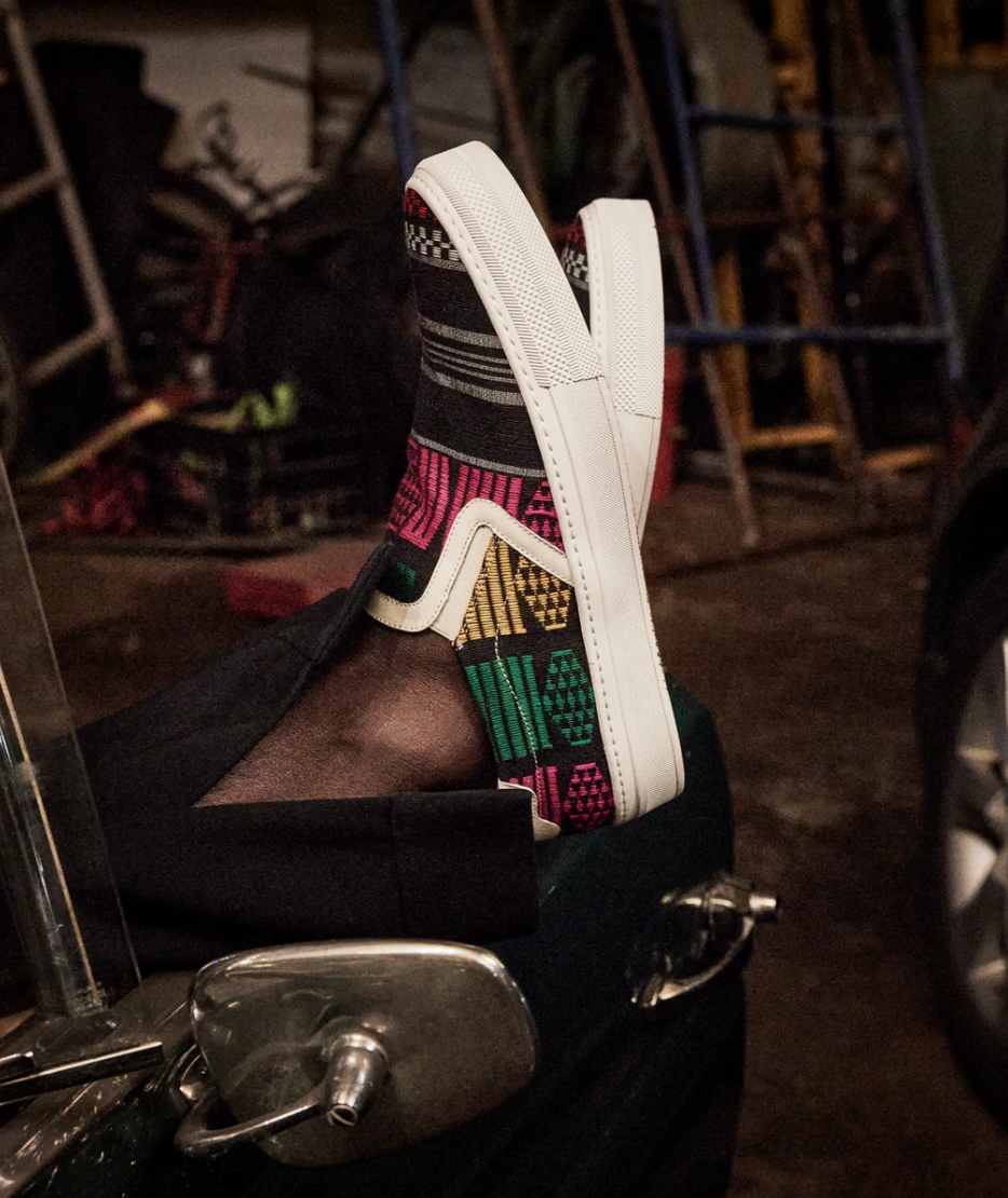 a mans feet wearing African pattered slip on shoes, propped up on a vintage car door 