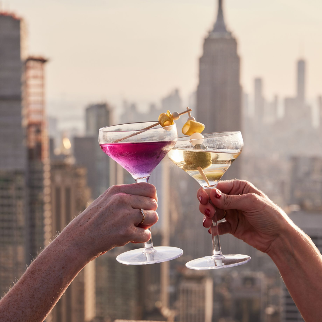 Two hands clinking cocktail glasses on The Weather Room's outdoor terrace at Top of the Rock