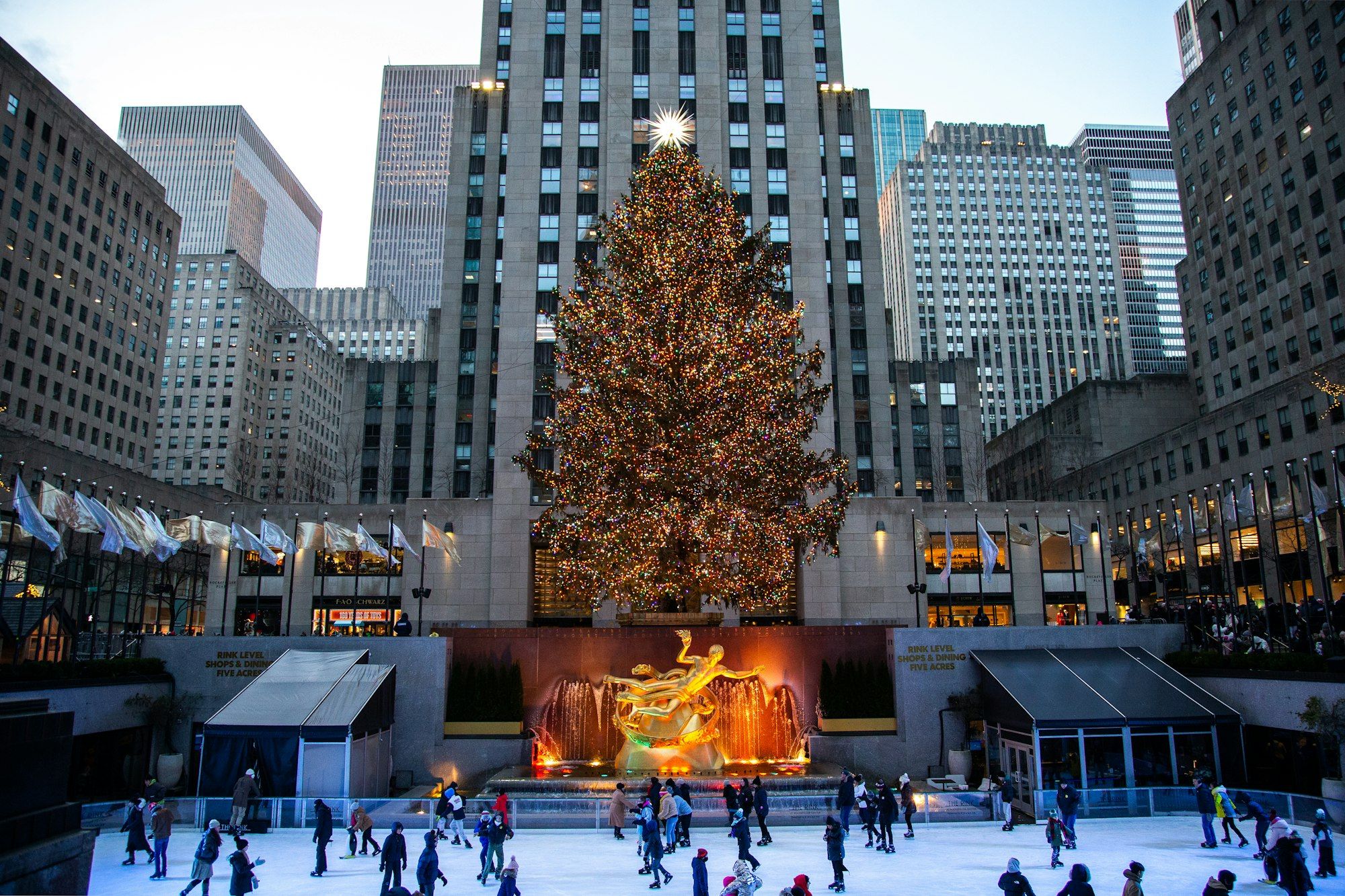 The Rink at Rockefeller Center glowing at dusk, with skaters on the ice beneath the Prometheus statue and the Christmas Tree.