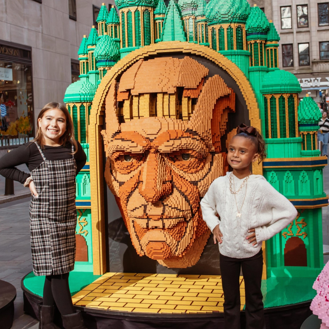Two children posing for a photo in front of a 'Wicked'-themed LEGO installation at Rockefeller Center