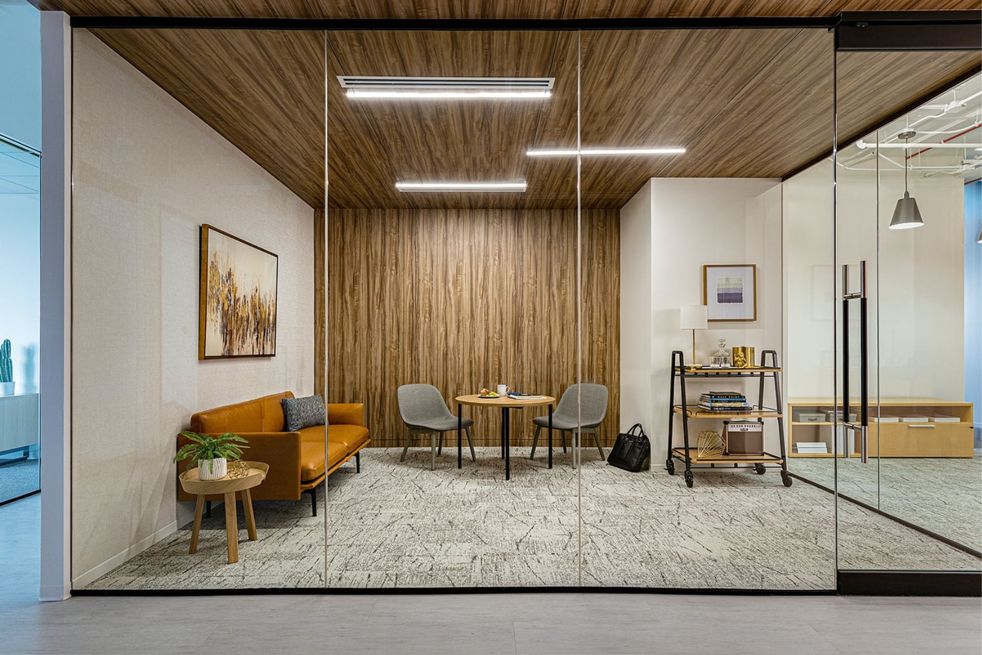 A modern office with a glass wall, wooden ceiling, and orange sofa, featuring a coffee table, plant, and desk, is illuminated by natural light and surrounded by a glass partition.