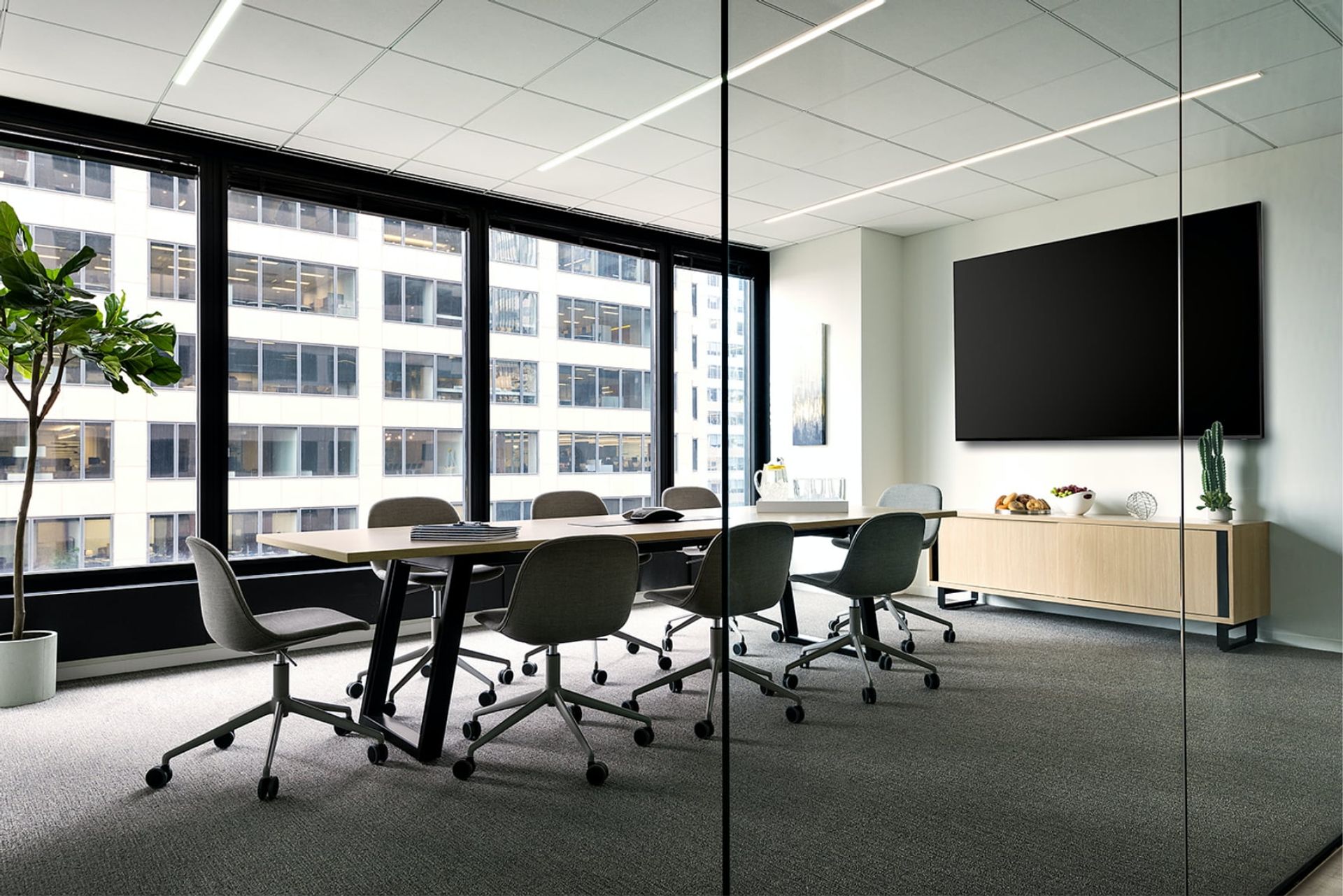 A modern office with a glass wall, large windows, and a desk with chairs, a potted plant, and a mounted television, creates a spacious and functional workspace.