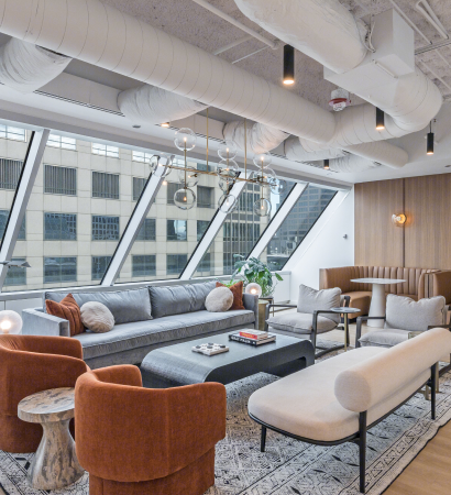 23rd floor clubhouse lounge at 525 West Monroe with floor‑to‑ceiling angled windows, a gray sofa, modern armchairs, a light bench, and a booth seating area under exposed ceiling ducts.