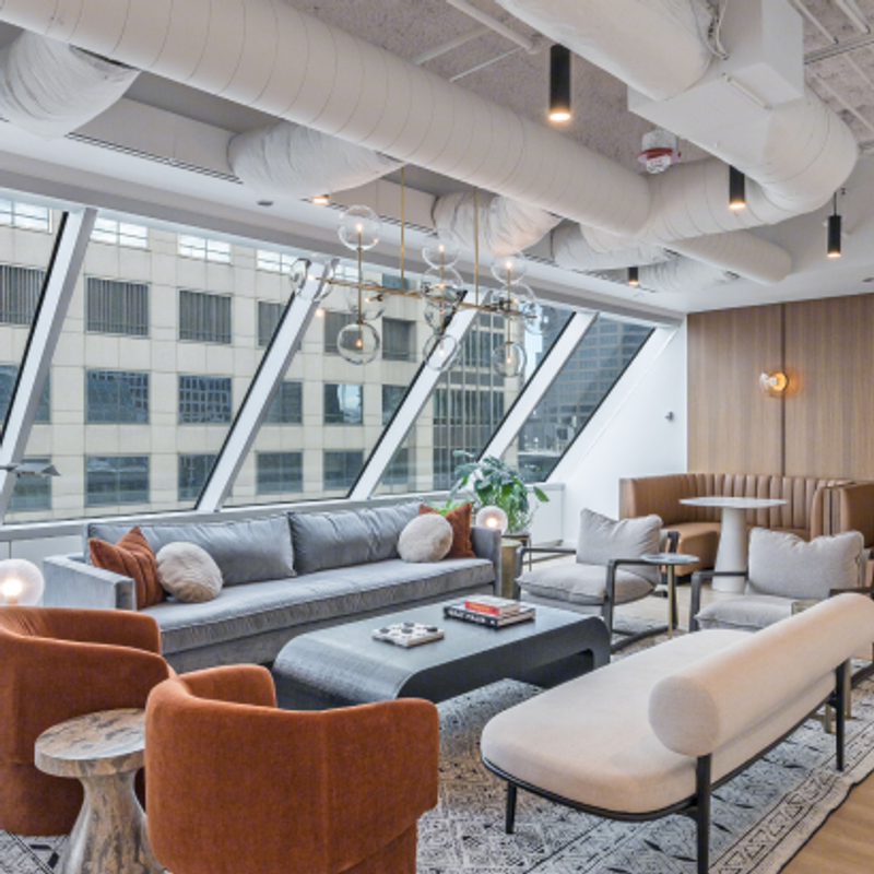 23rd floor clubhouse lounge at 525 West Monroe with floor‑to‑ceiling angled windows, a gray sofa, modern armchairs, a light bench, and a booth seating area under exposed ceiling ducts.
