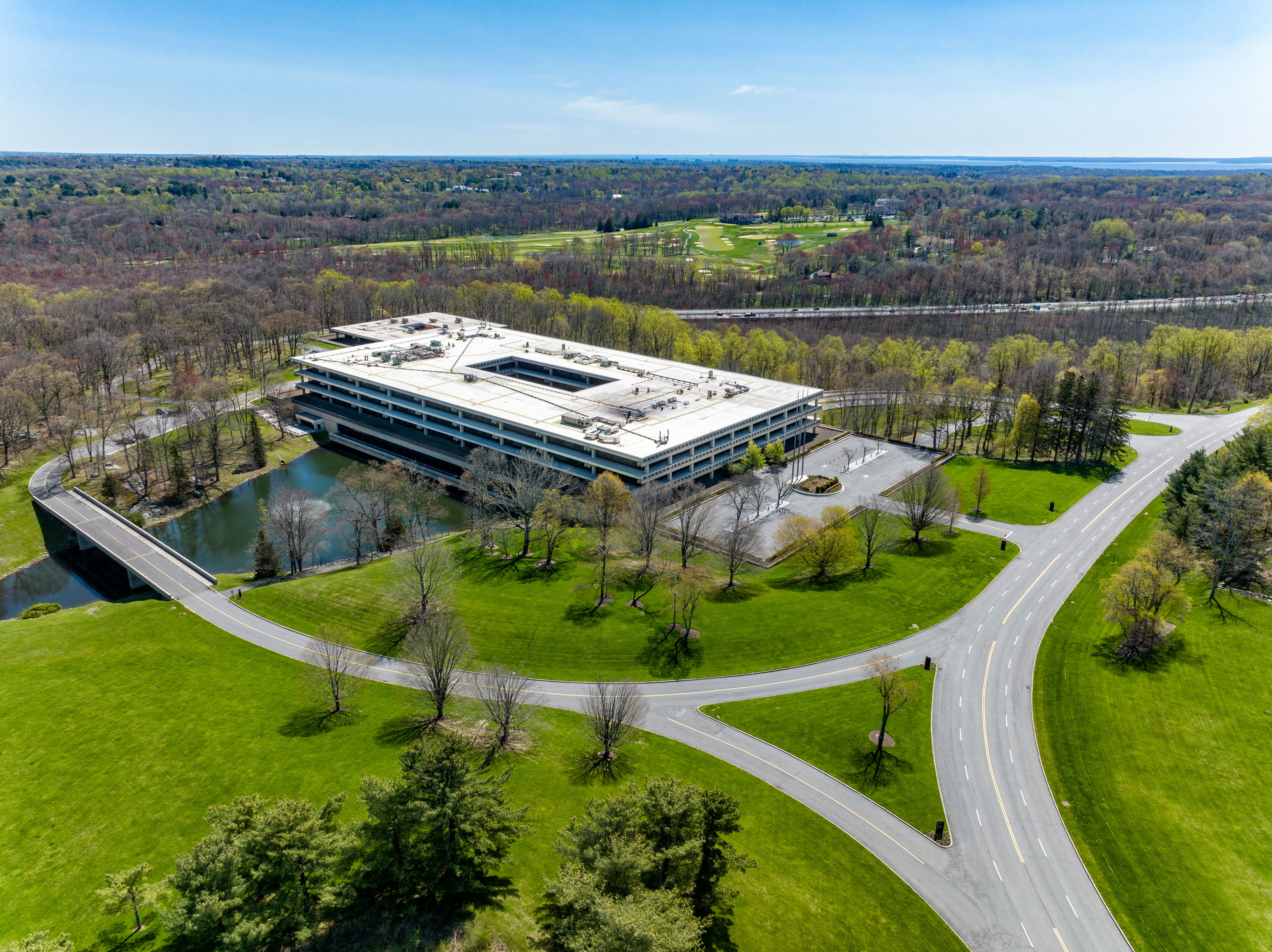 An aerial view of a modern, large building surrounded by greenery and a pond, with a road and parking lot nearby.