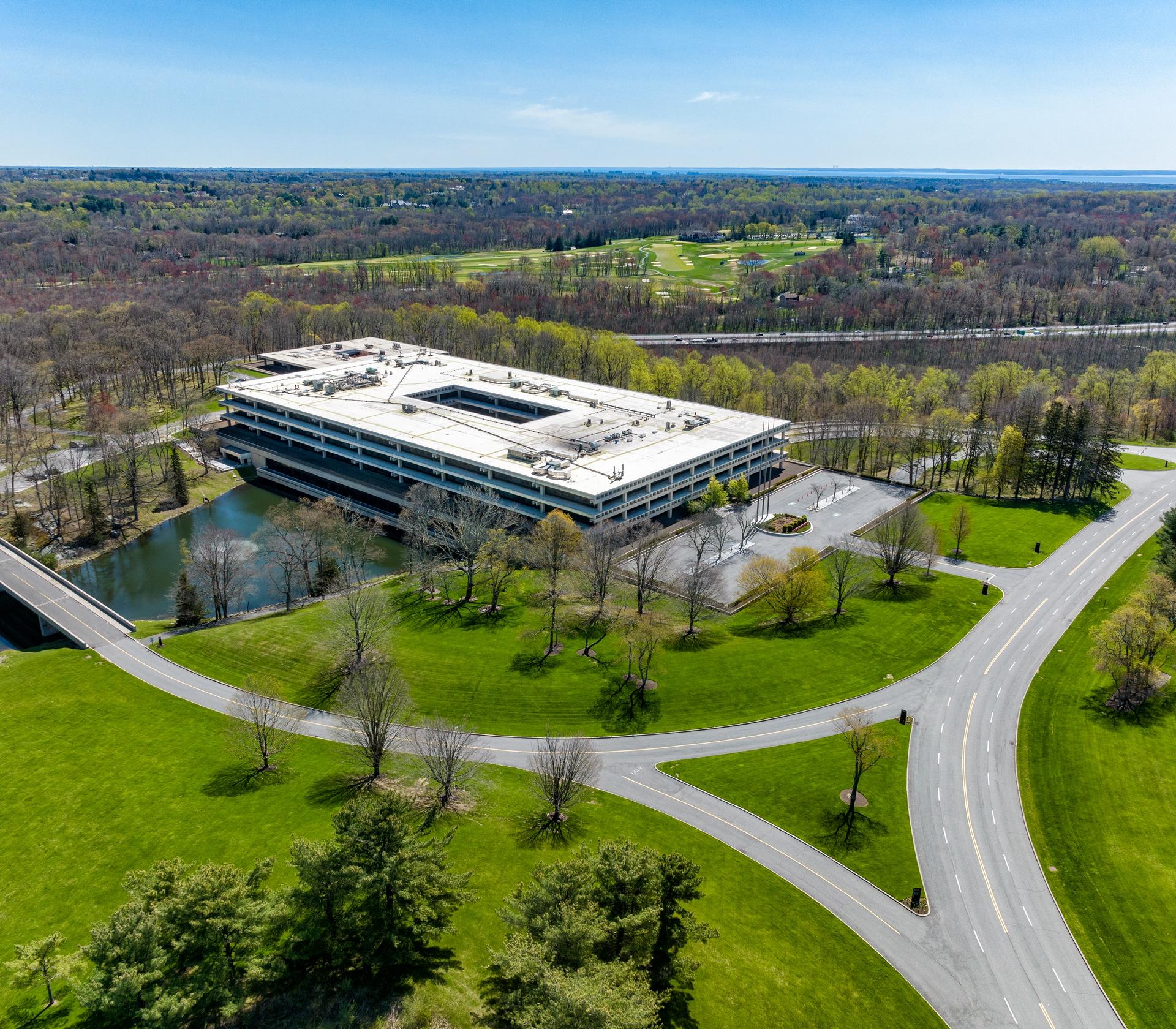 An aerial view of a modern, large building surrounded by greenery and a pond, with a road and parking lot nearby.
