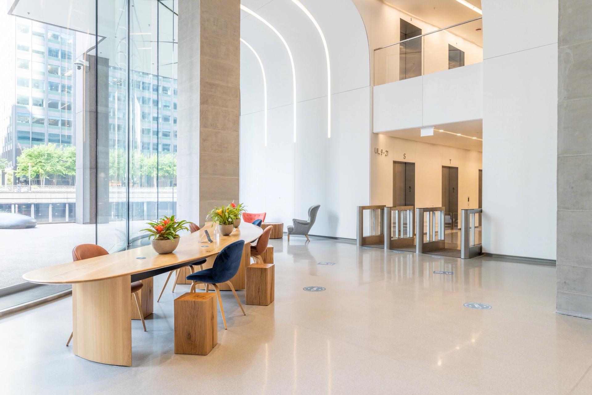 A modern office space with a large glass window, white marble floor, and a wooden table with flowers, surrounded by modern furniture and a glass-enclosed elevator.