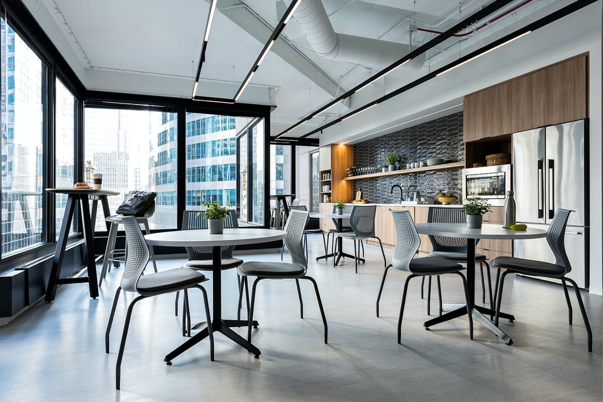 A modern office space with a large window overlooking a city skyline, featuring a modern kitchen, dining area, and a bar area with a barstool and potted plants.