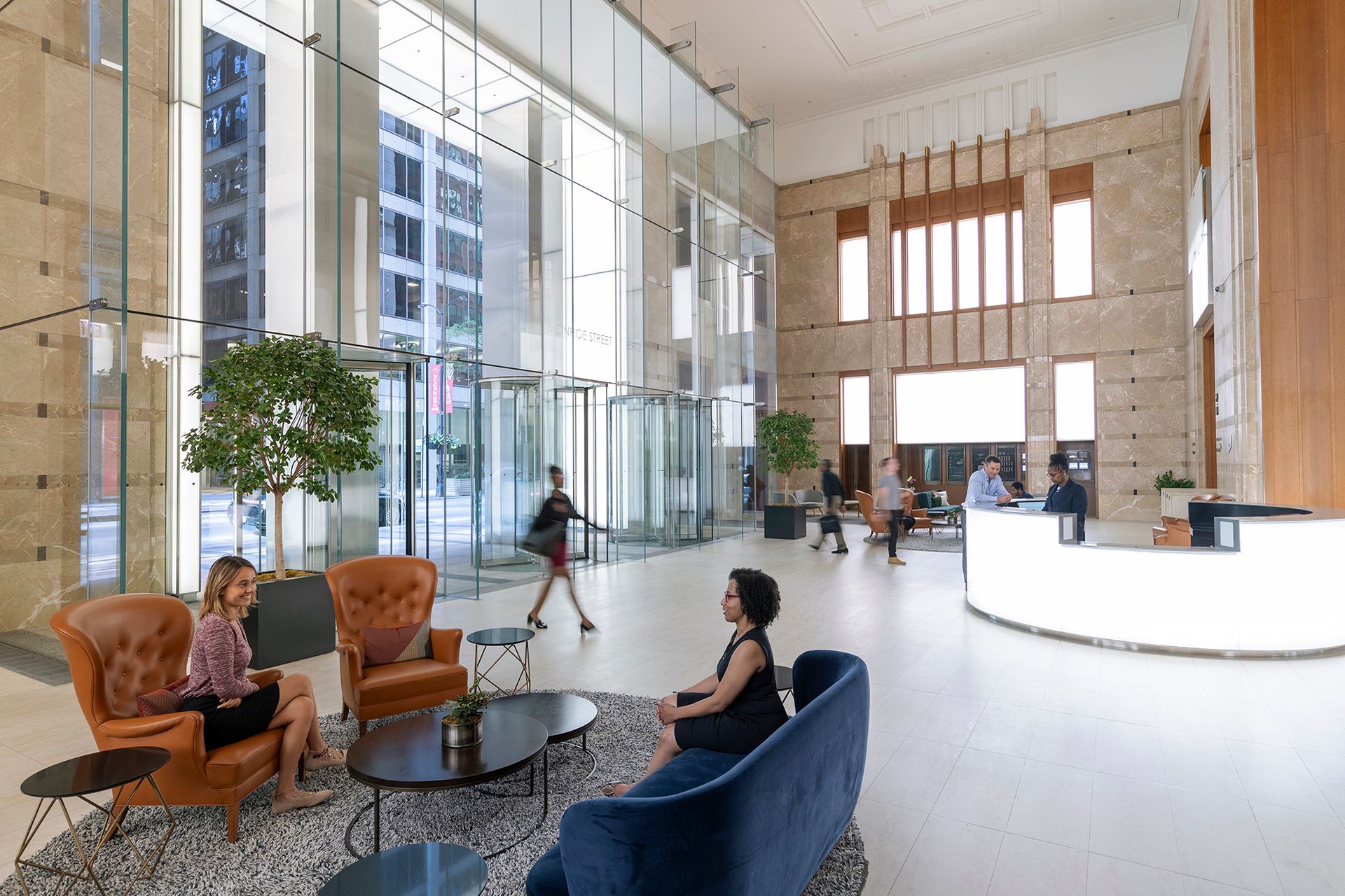 Spacious office lobby with people sitting and walking, featuring large glass windows and a reception desk