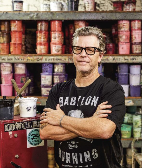 Adam Savage in a workshop filled with labeled containers, sporting his signature thick-rimmed glasses