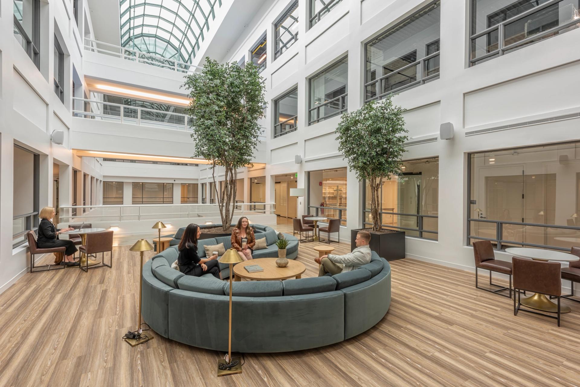 A modern indoor lounge area with curved sofas, round tables, chairs, and several people working or relaxing under natural light and greenery.