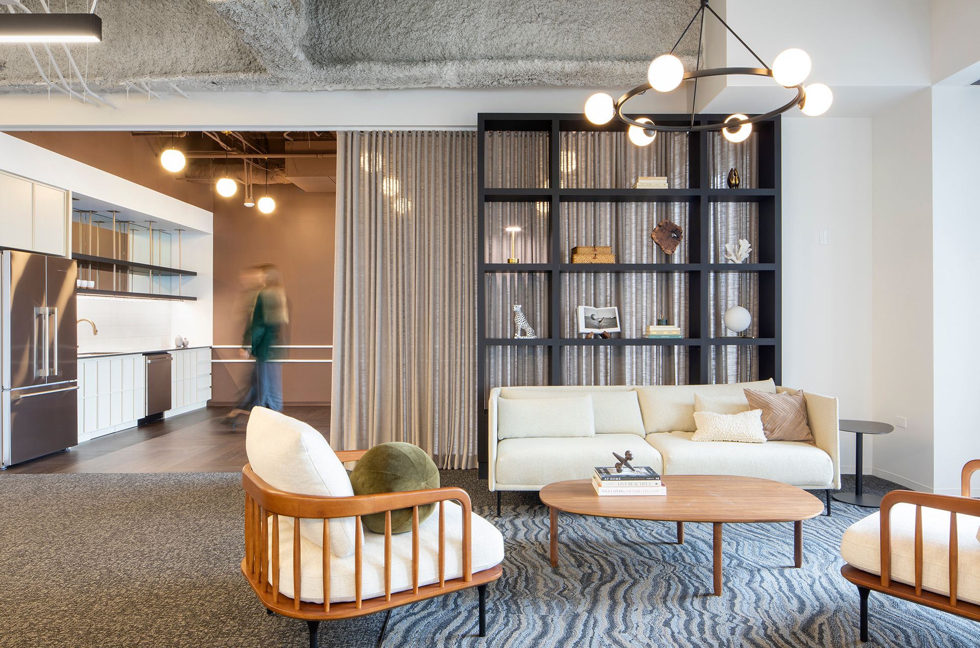 Modern living room with beige sofa, wooden armchairs, coffee table, black shelving unit, and an open kitchen area in the background