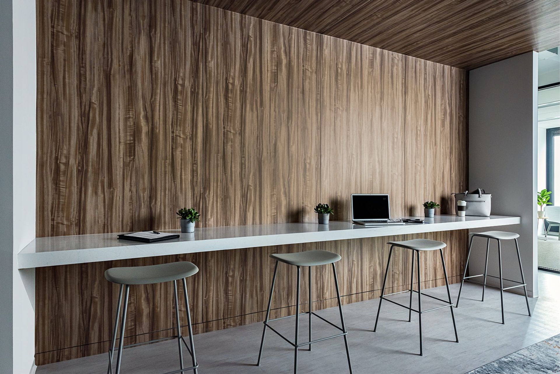 A modern office features a long white counter with a laptop, stools, and potted plants, creating a functional and stylish workspace.