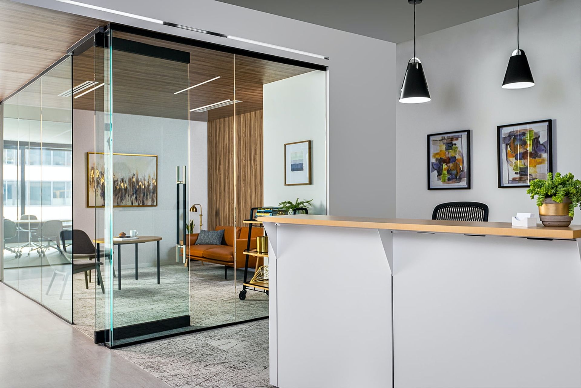 A modern office features a glass wall, wooden desk, and potted plant, with a glass door leading to a balcony and a wooden door leading to a second floor.
