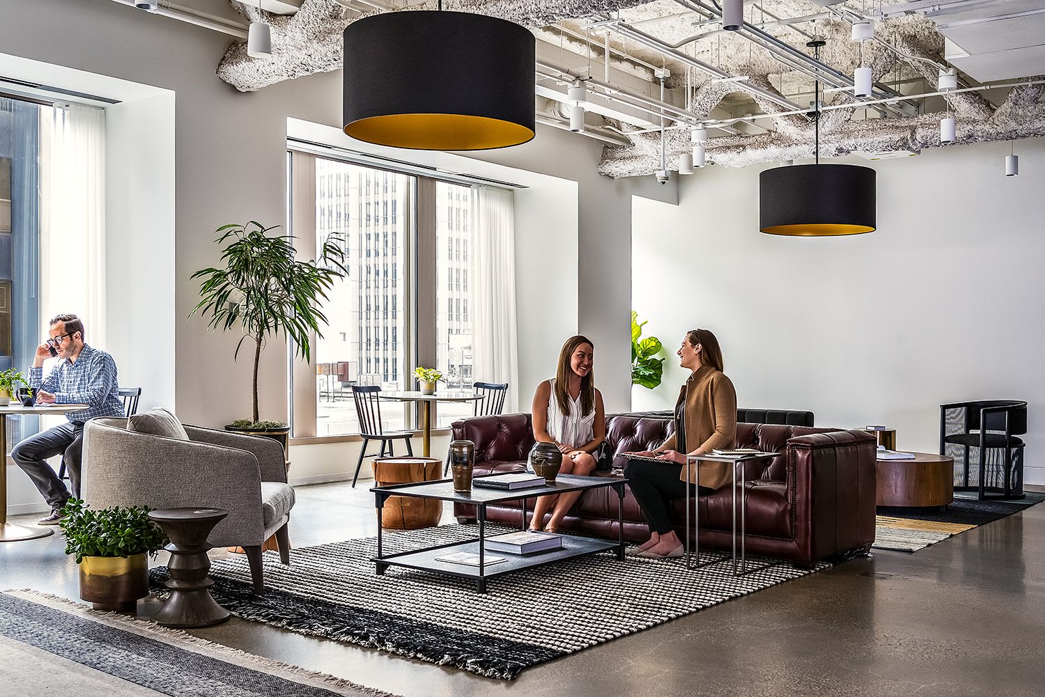 Two women sitting and talking on a brown leather sofa in a modern office lounge with large windows and stylish decor