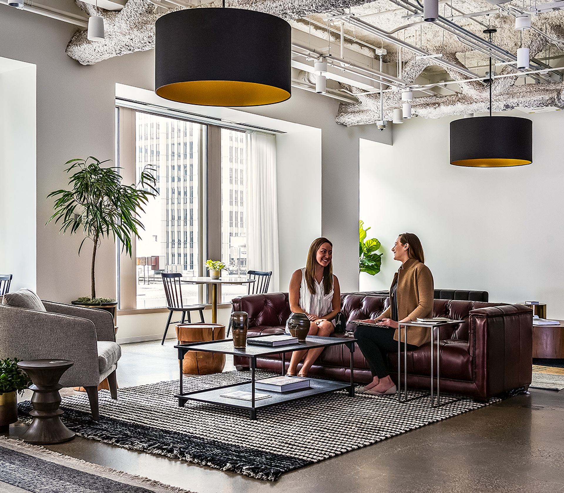 Two women sitting and talking on a brown leather sofa in a modern office lounge with large windows and stylish decor