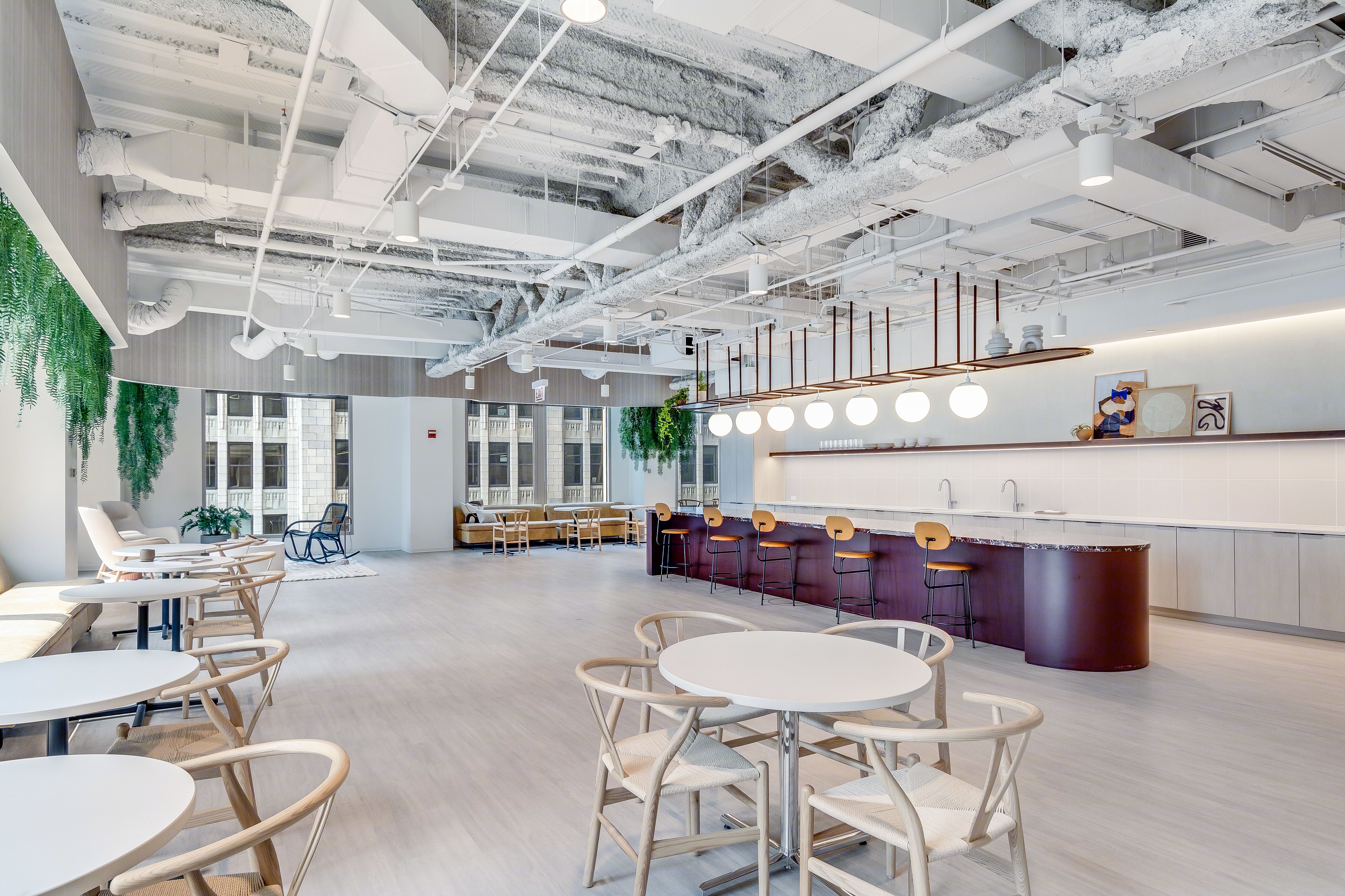 Bright and modern open office lounge space with round tables, wooden chairs, bar stools, and hanging plants