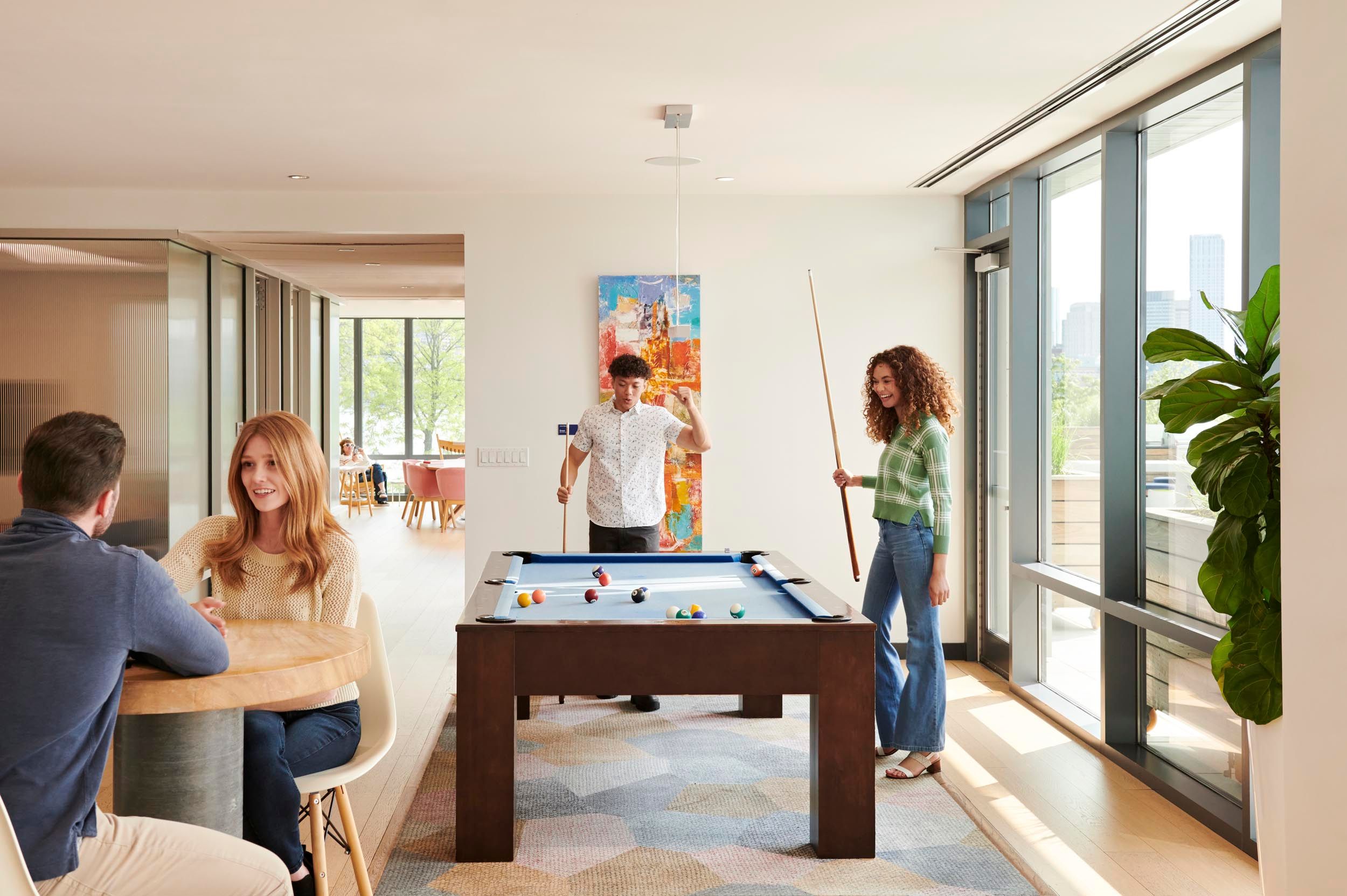 Resident lounge at The Eddy Apartments for rent in East Boston, featuring floor to ceiling windows and people playing pool.