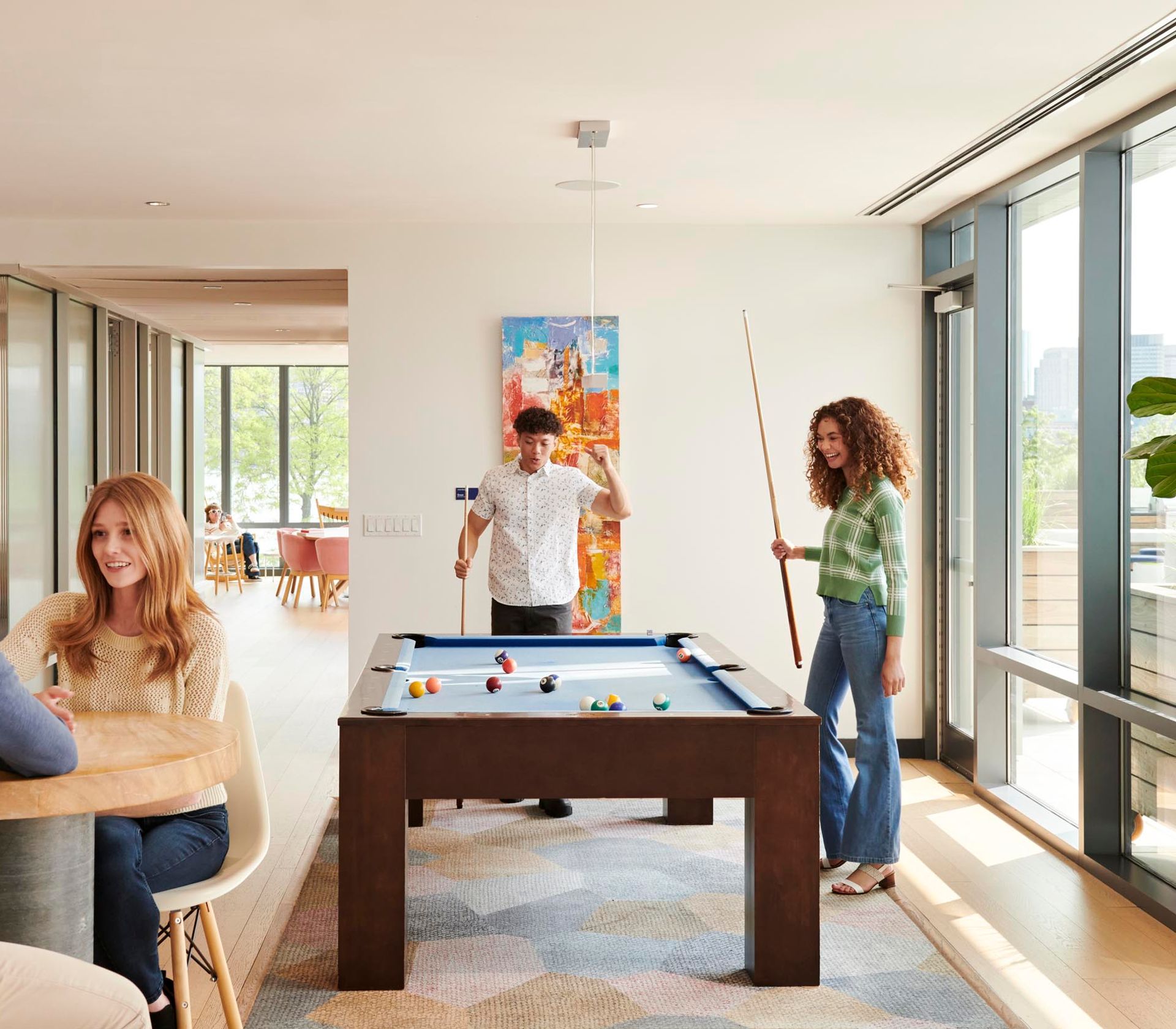 Resident lounge at The Eddy Apartments for rent in East Boston, featuring floor to ceiling windows and people playing pool.