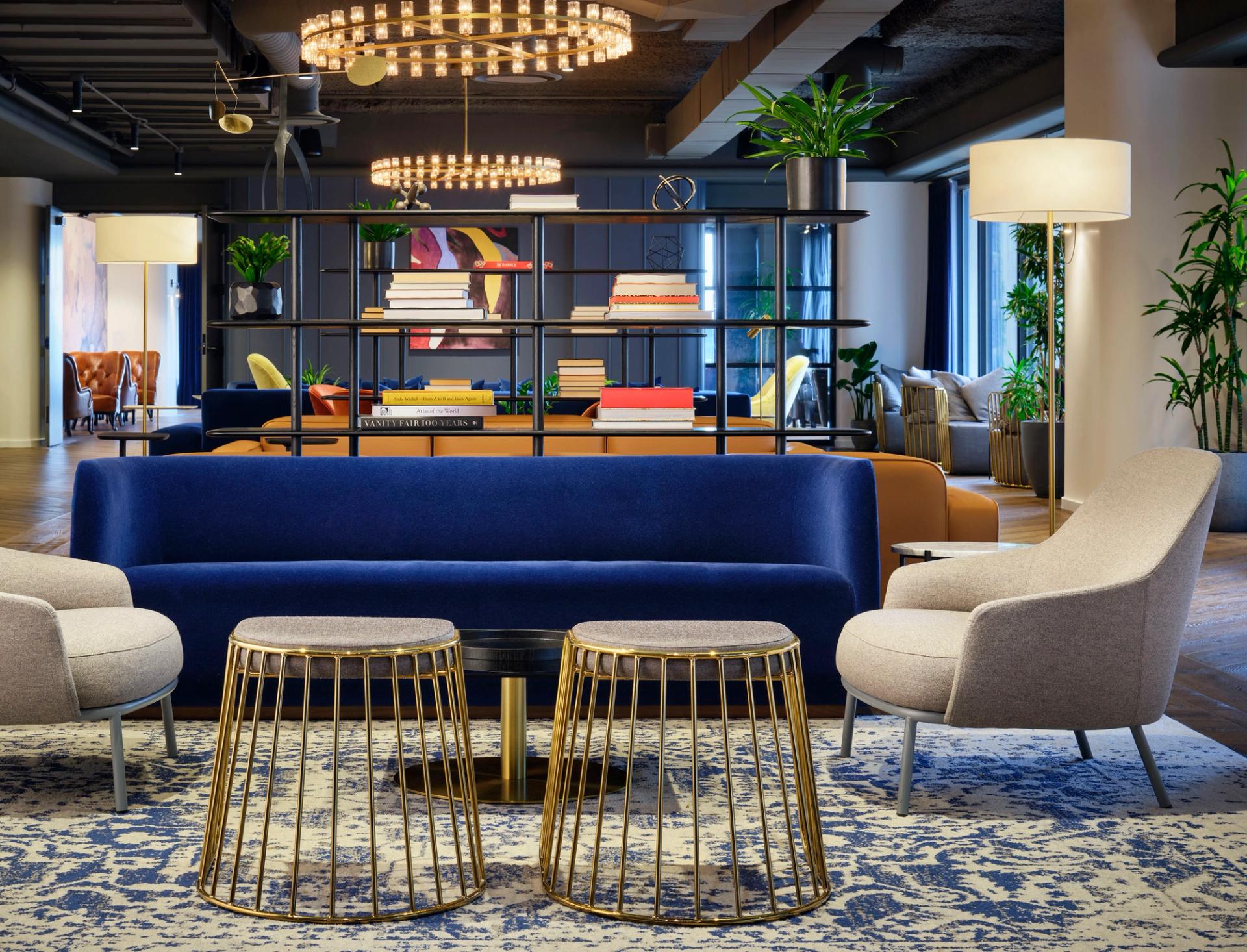 This modern lounge interior features a bold blue velvet sofa paired with gold wire-frame coffee tables and neutral-toned armchairs under a high industrial ceiling.