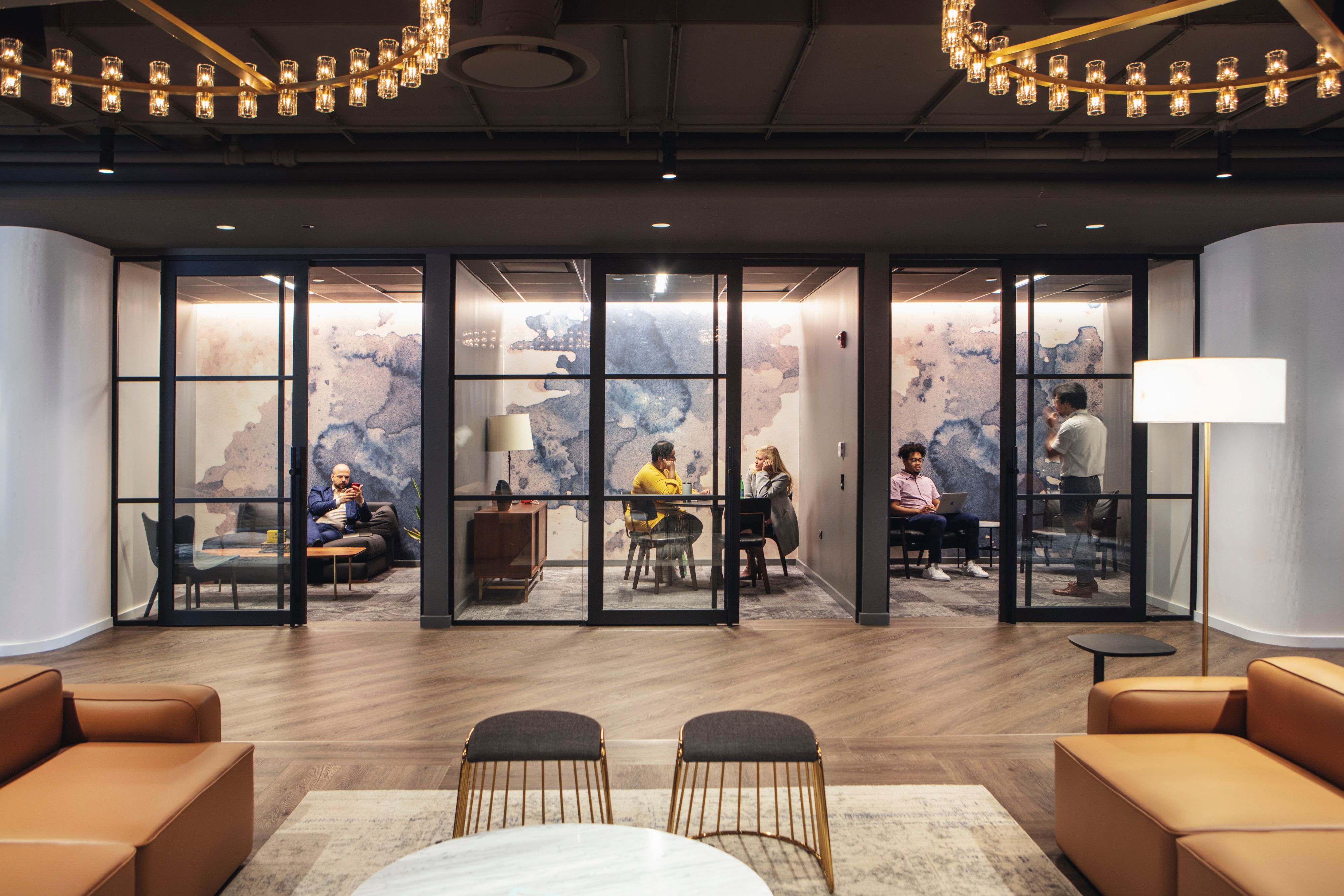 The modern office space features a large glass wall, a chandelier, and a cozy seating area with leather sofas and chairs, surrounded by a coffee table and a bar area.
