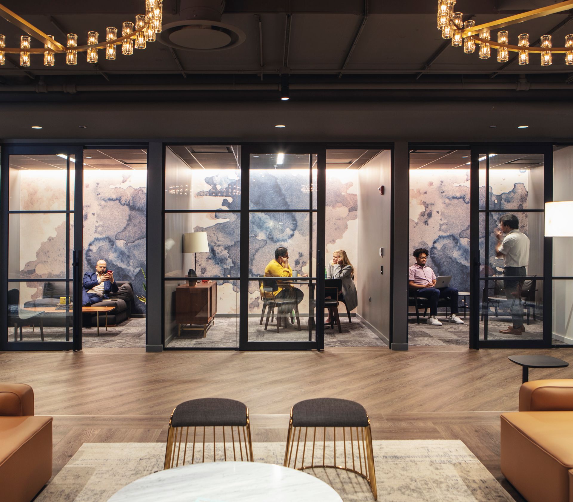 The modern office space features a large glass wall, a chandelier, and a cozy seating area with leather sofas and chairs, surrounded by a coffee table and a bar area.