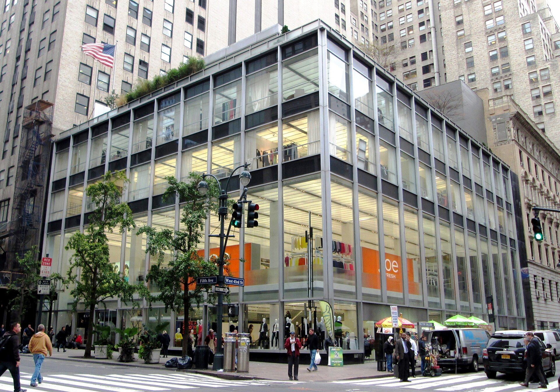 A bustling city street features a large glass building with an orange banner, a pedestrian crossing, a parked van, and a street vendor, while tall buildings and a tall flag add to the urban atmosphere.
