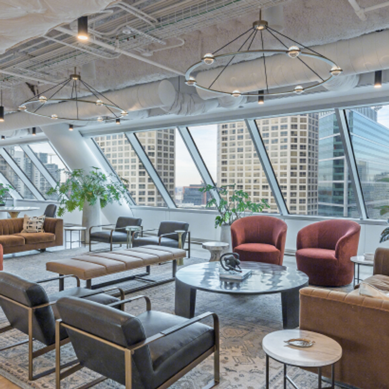 23rd floor clubhouse lounge at 525 West Monroe with floor-to-ceiling angled windows, leather and fabric seating in warm tones, round coffee tables, greenery, and modern circular chandeliers.