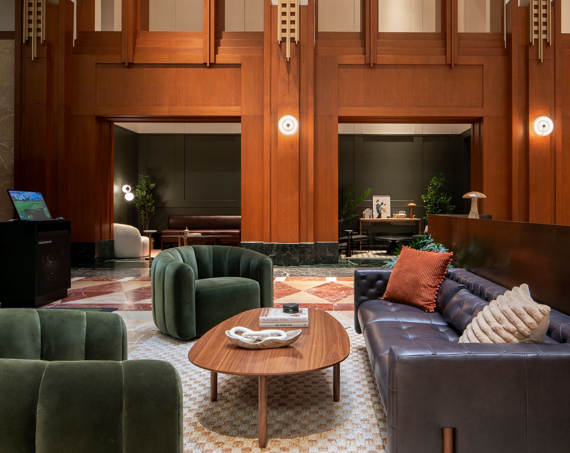 Modern lounge area with green velvet chairs, a leather sofa with pillows, and a wooden coffee table in front of wood-paneled walls