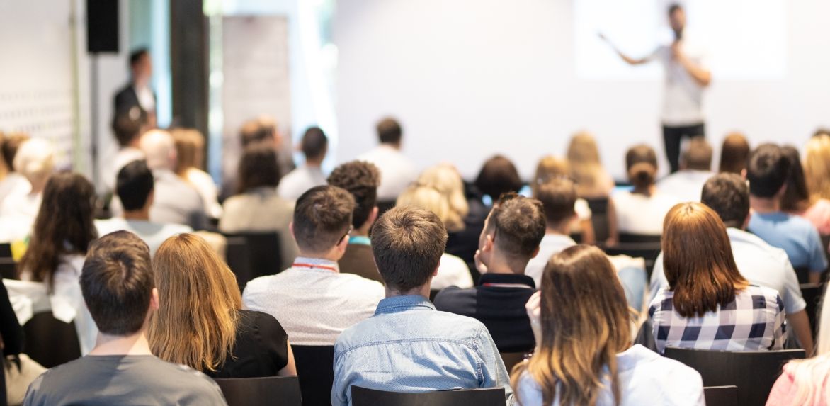 Audience of people seated and facing a presentation screen in a conference room