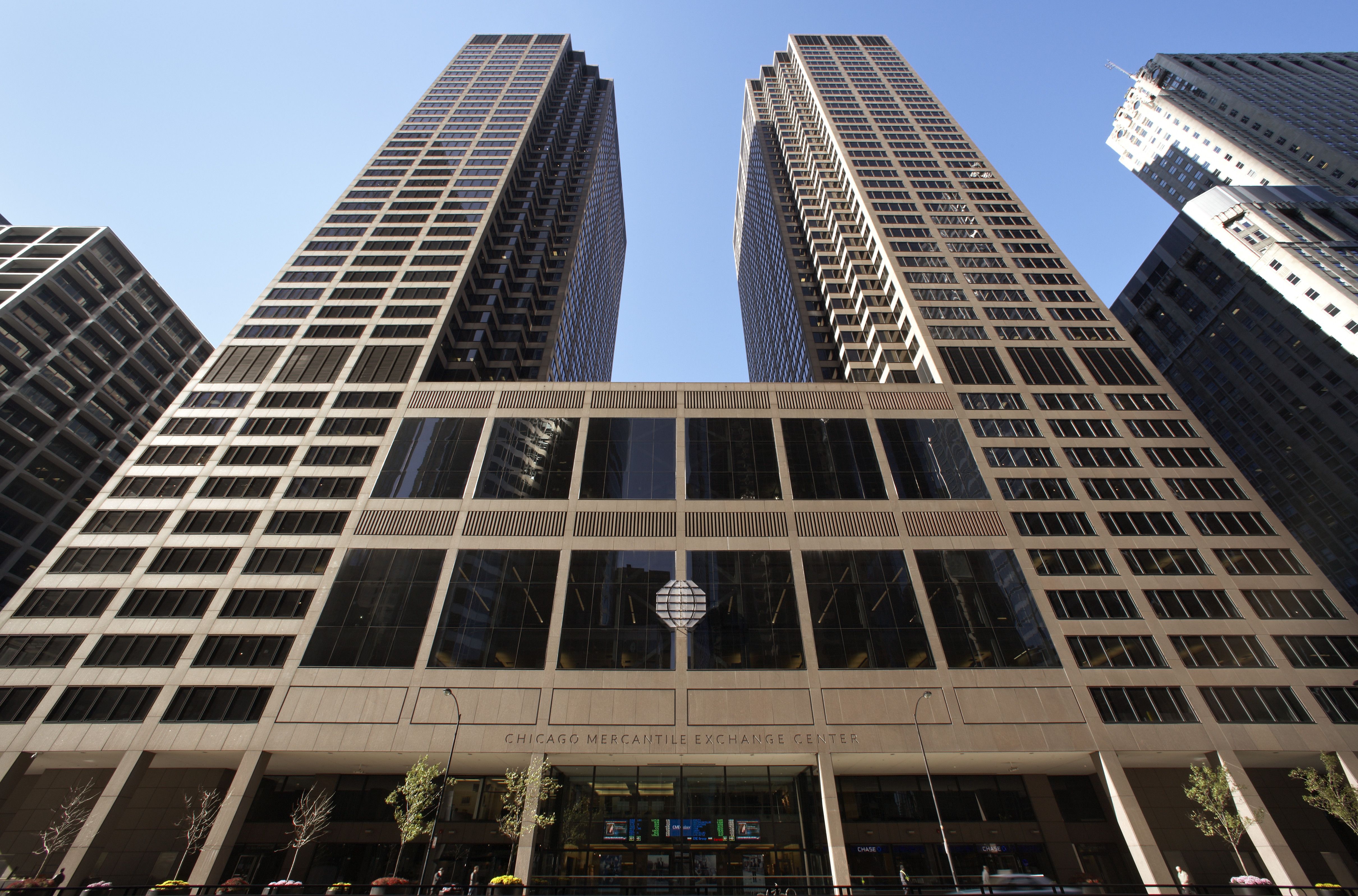 Two tall, modern skyscrapers with large glass windows stand side by side in a bustling cityscape, surrounded by parked cars and a clear blue sky.