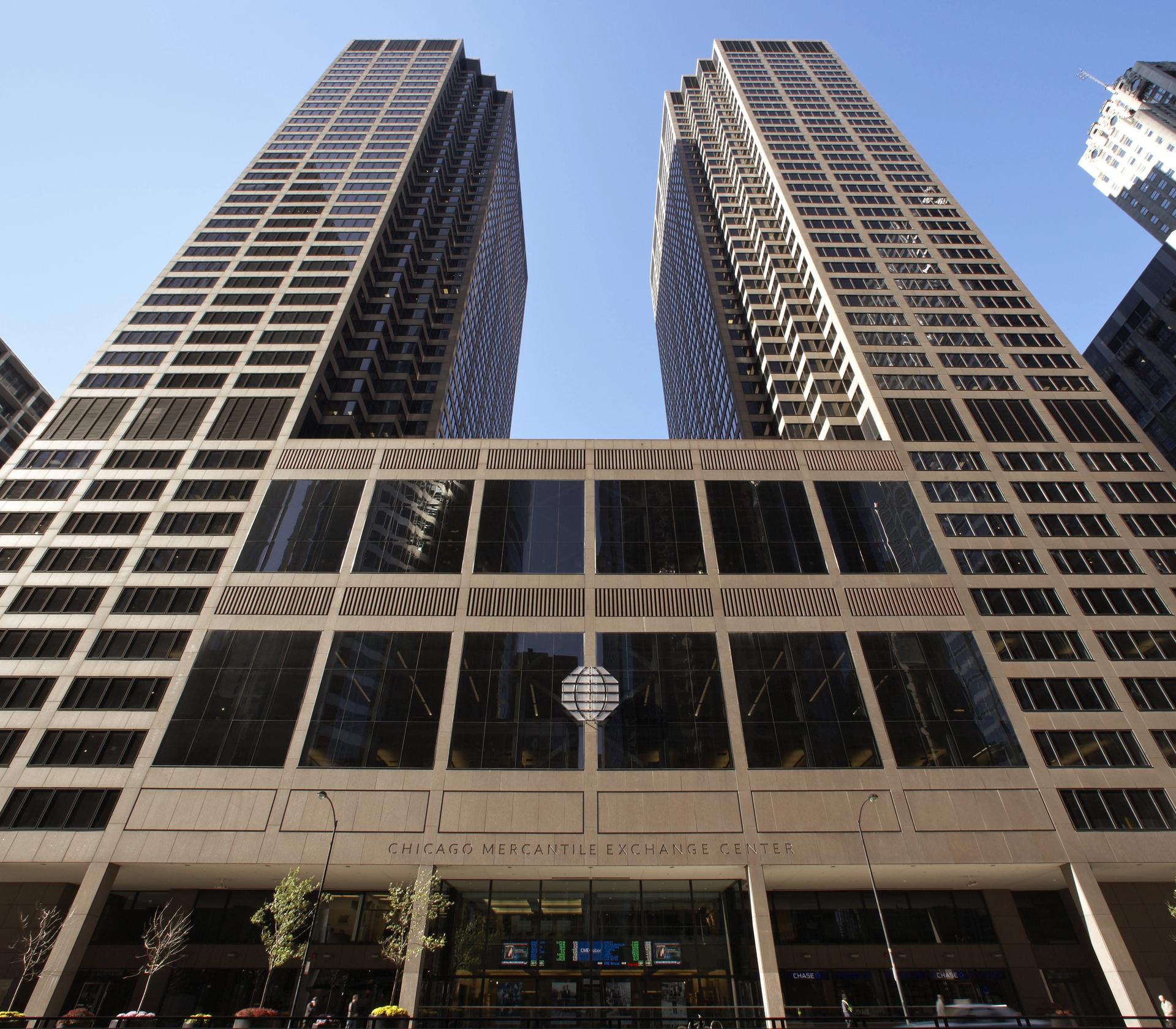 Two tall, modern skyscrapers with large glass windows stand side by side in a bustling cityscape, surrounded by parked cars and a clear blue sky.