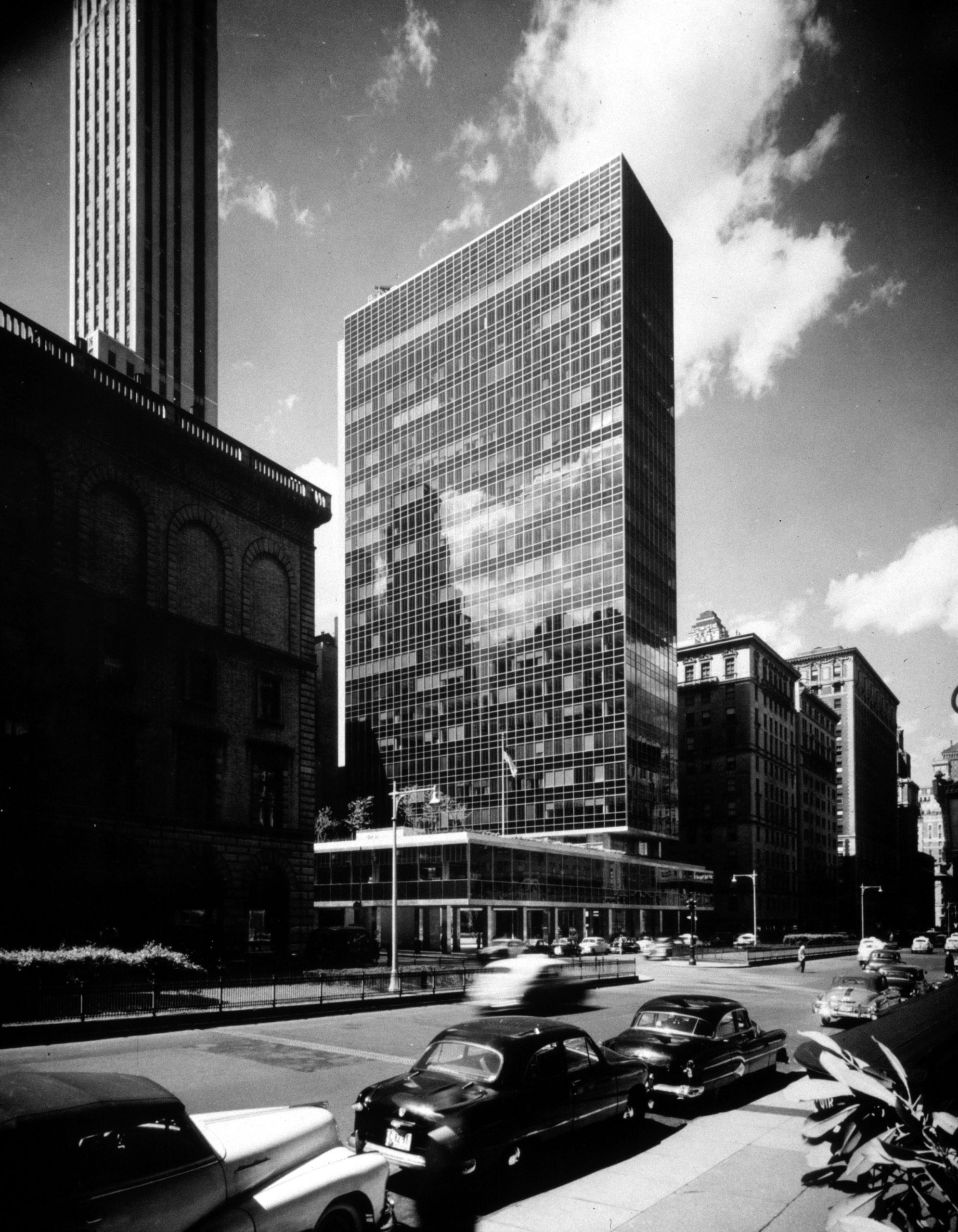 A bustling city street features a large glass building, a tall brick building, and a smaller building, with cars driving on the road and a pedestrian crossing the street.