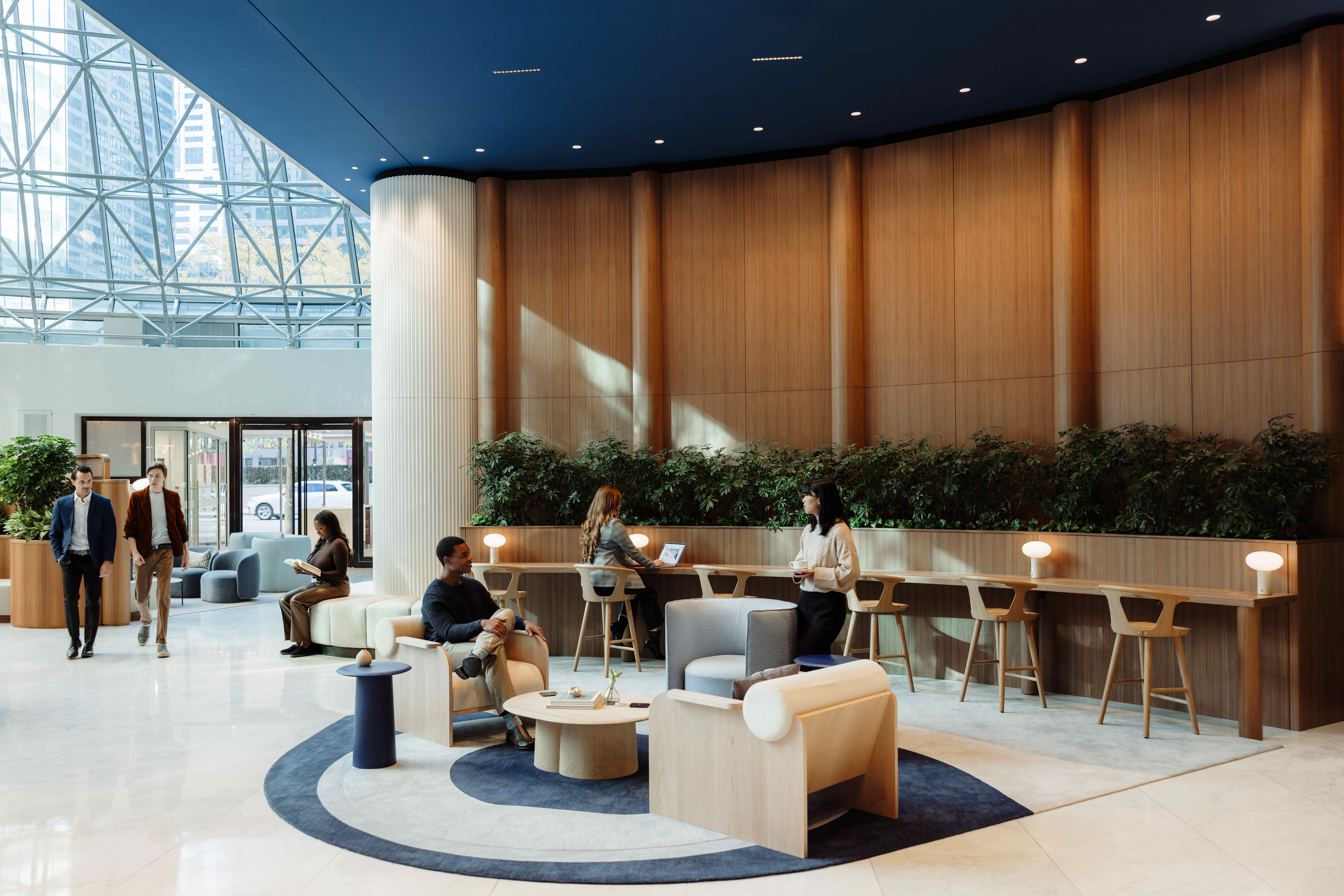 Lobby lounge at 525 West Monroe with modern seating, circular rugs, tall wooden panel walls, built-in planter greenery, and people working on laptops under natural light from the glass atrium ceiling.